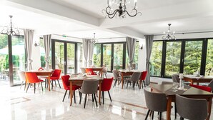 A spacious dining area with multiple tables and chairs, large windows with curtains, and a chandelier.