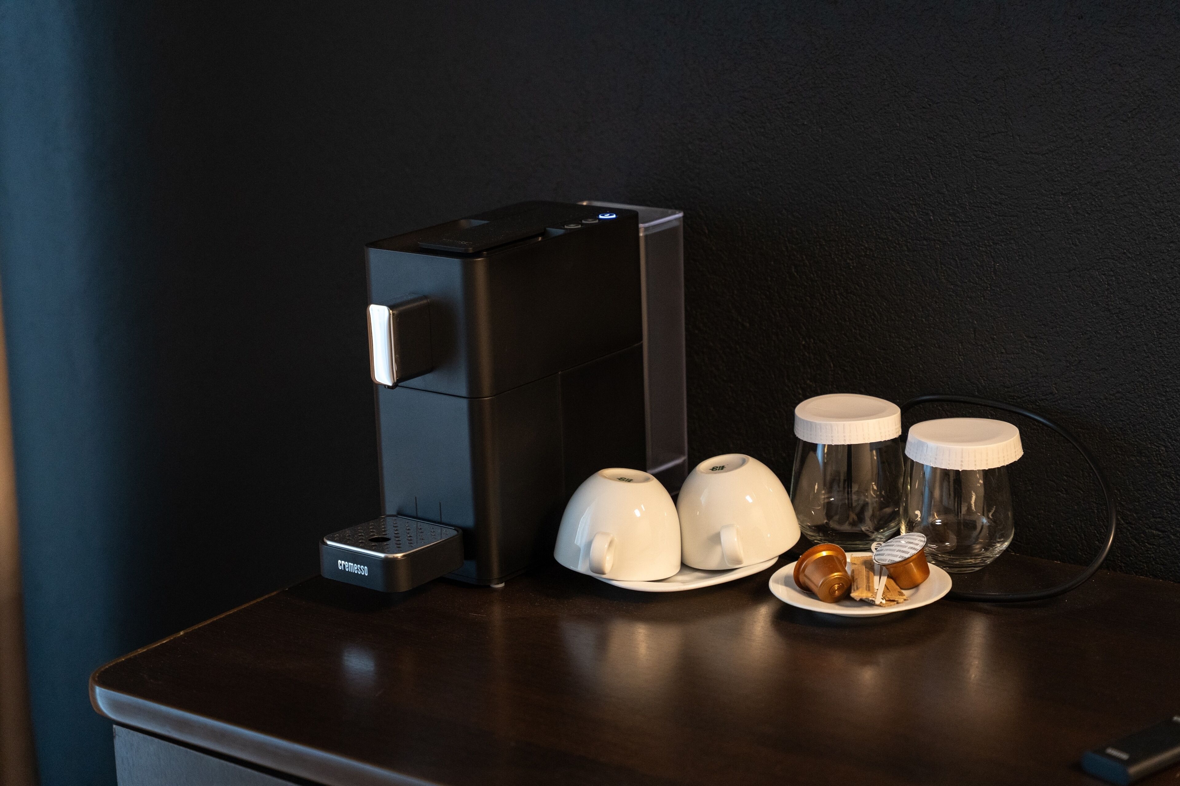A coffee maker, two white cups, and two glass jars with lids on a dark wooden table.