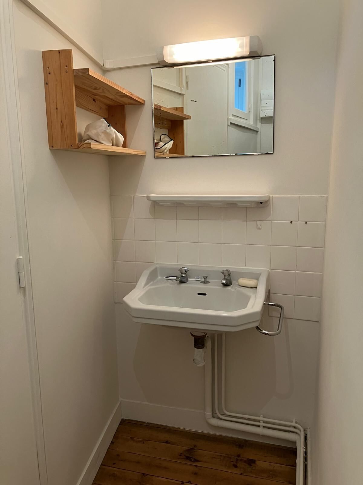 Ein kleines Badezimmer mit einem weißen Waschbecken, Holzregalen, einem Spiegel und weiß gefliesten Wänden.