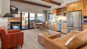 A modern living room with a fireplace, a sofa, a coffee table, and a kitchen area with wooden cabinets and a stainless steel refrigerator. - Newpark 315 | Private Hot Tub | Kitchenette | Mountain & Preserve Views | Fireplace | Washer/Dryer (Park City)
