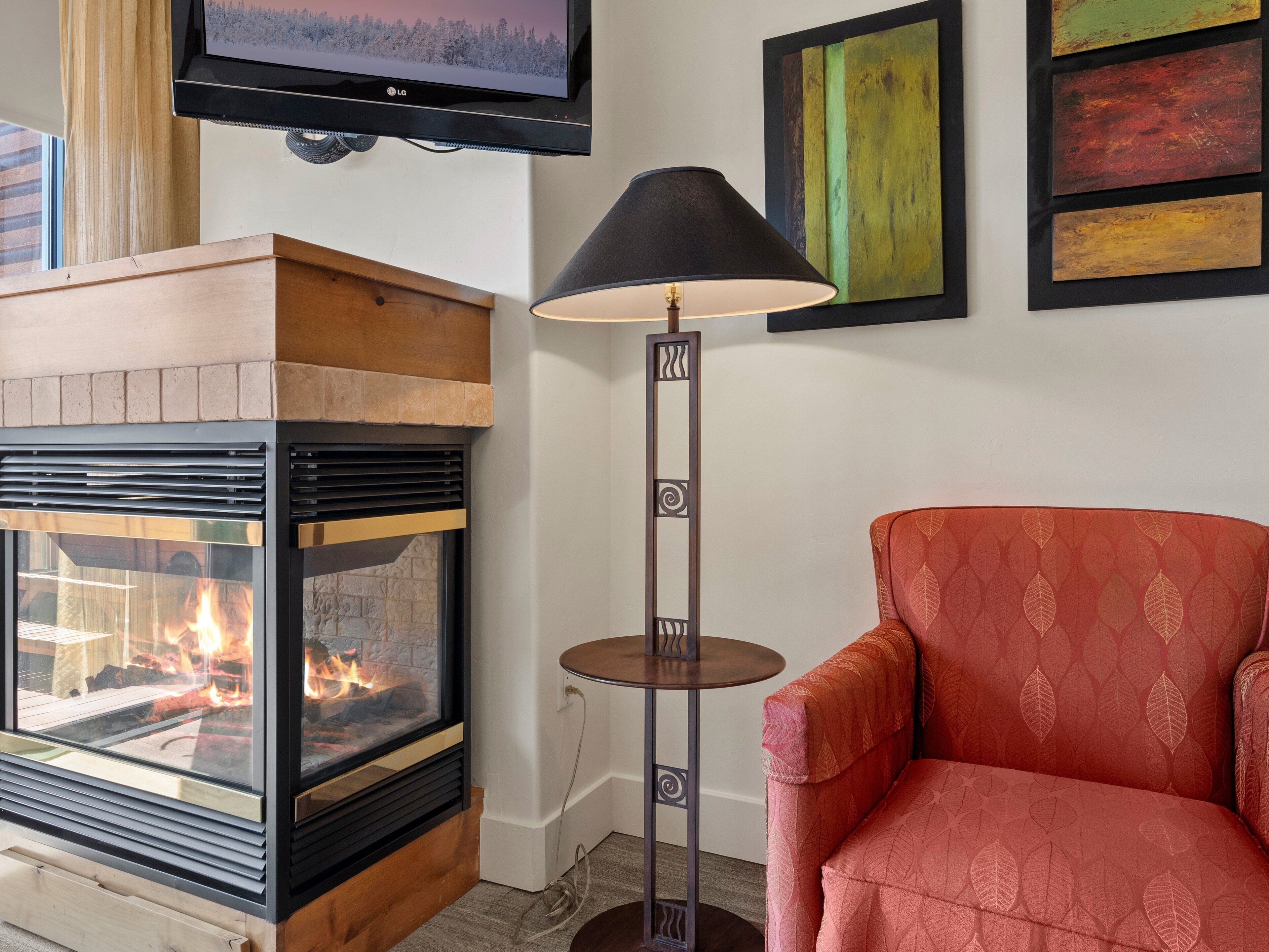 A cozy living room with a fireplace, a comfortable red armchair, a floor lamp, and a flat-screen TV mounted above the fireplace.