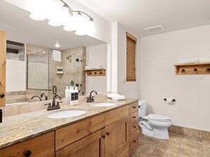 Een moderne badkamer met twee wasbakken, een douche en een toilet. Er zijn houten kastjes en een grote spiegel boven de wasbakken.