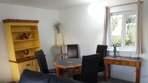 A room with a dining table, chairs, a sofa, a wooden cabinet, and a window with a view of greenery. - Thistle Cottage (Ryde)