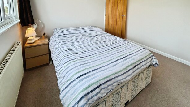 A bedroom with a bed, a nightstand, a lamp, a window with curtains, and a door. - Beachfield Breeze (Bembridge)
