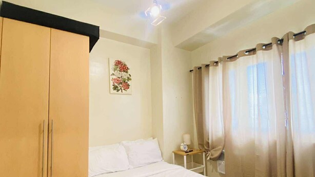 A bedroom with a bed, a wardrobe, a small table with a lamp, and a wall decoration. - Room-cozy Iloilo City Condo Room With Sh (Iloilo)
