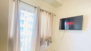 A hotel room with a bed, a flat-screen TV mounted on the wall, a window with curtains, and a view of a building outside. - Room-cozy Iloilo City Condo Room With Sh (Iloilo)
