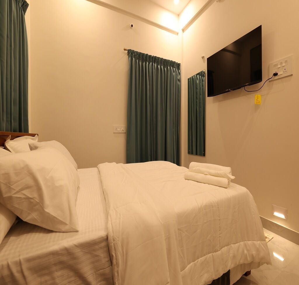 A neatly made bed with white linens, a flat-screen TV mounted on the wall, and a window with green curtains.