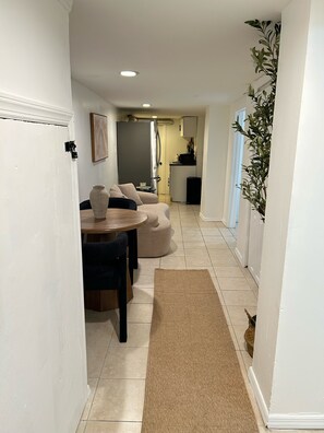 A narrow hallway leading to a dining area with a table and chairs, a sofa, and a kitchen area in the background. - AUREA Stays /Private 2BR Suite / Free Parking /Near EWR (Newark)