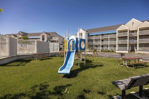 A playground with a slide, benches, and picnic tables in a grassy area with apartment buildings in the background.