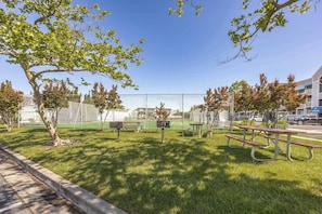 A park with picnic tables, a fenced tennis court, and trees.