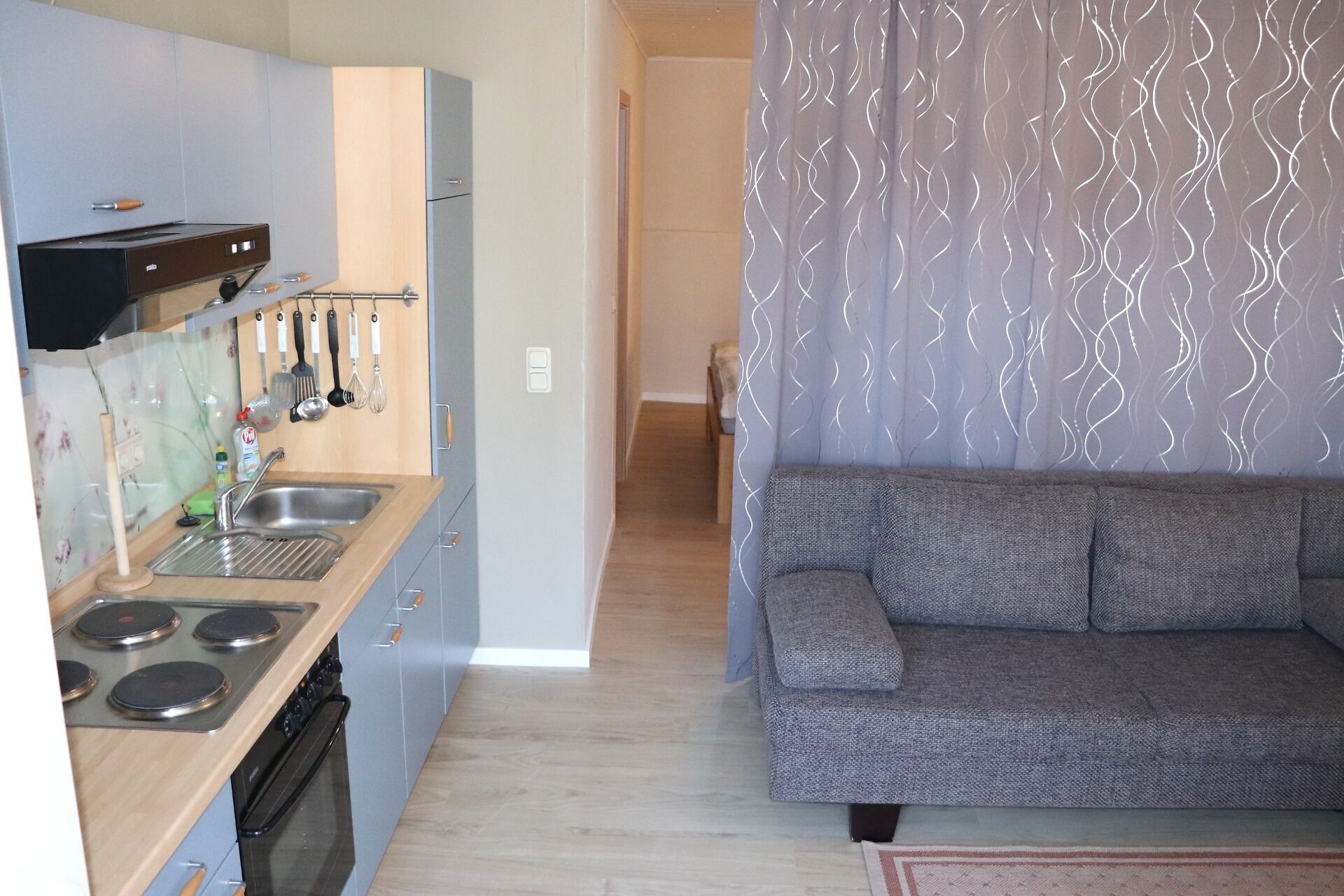 A compact kitchen with a stove, sink, and hanging utensils. A grey sofa is positioned in the living area.