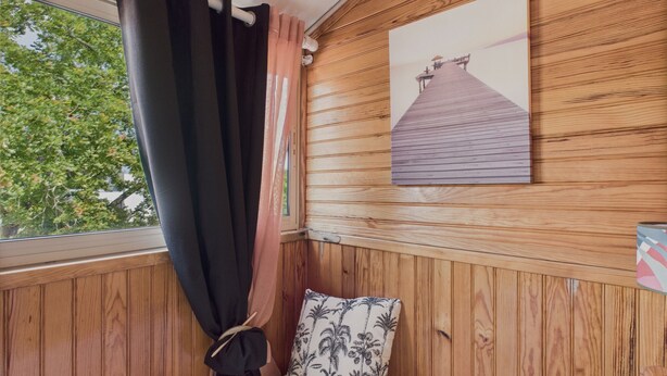 A wooden cabin room with a bed, a window with curtains, and a picture of a pier on the wall. - 300m from beach, Andernos centre – sleeps 4 (Andernos-les-Bains)