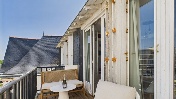 A wooden deck with a white table and bench, a bottle of wine, and a view of a building with a dark roof.
