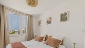 A bedroom with a large bed, two bedside tables, a view of the city through a window, and two framed pictures on the wall.