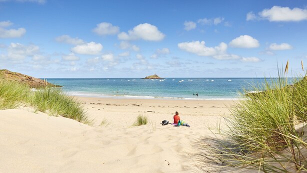Ein Strand mit einer Person, die im Sand sitzt, einer kleinen Insel in der Ferne und dem Meer.