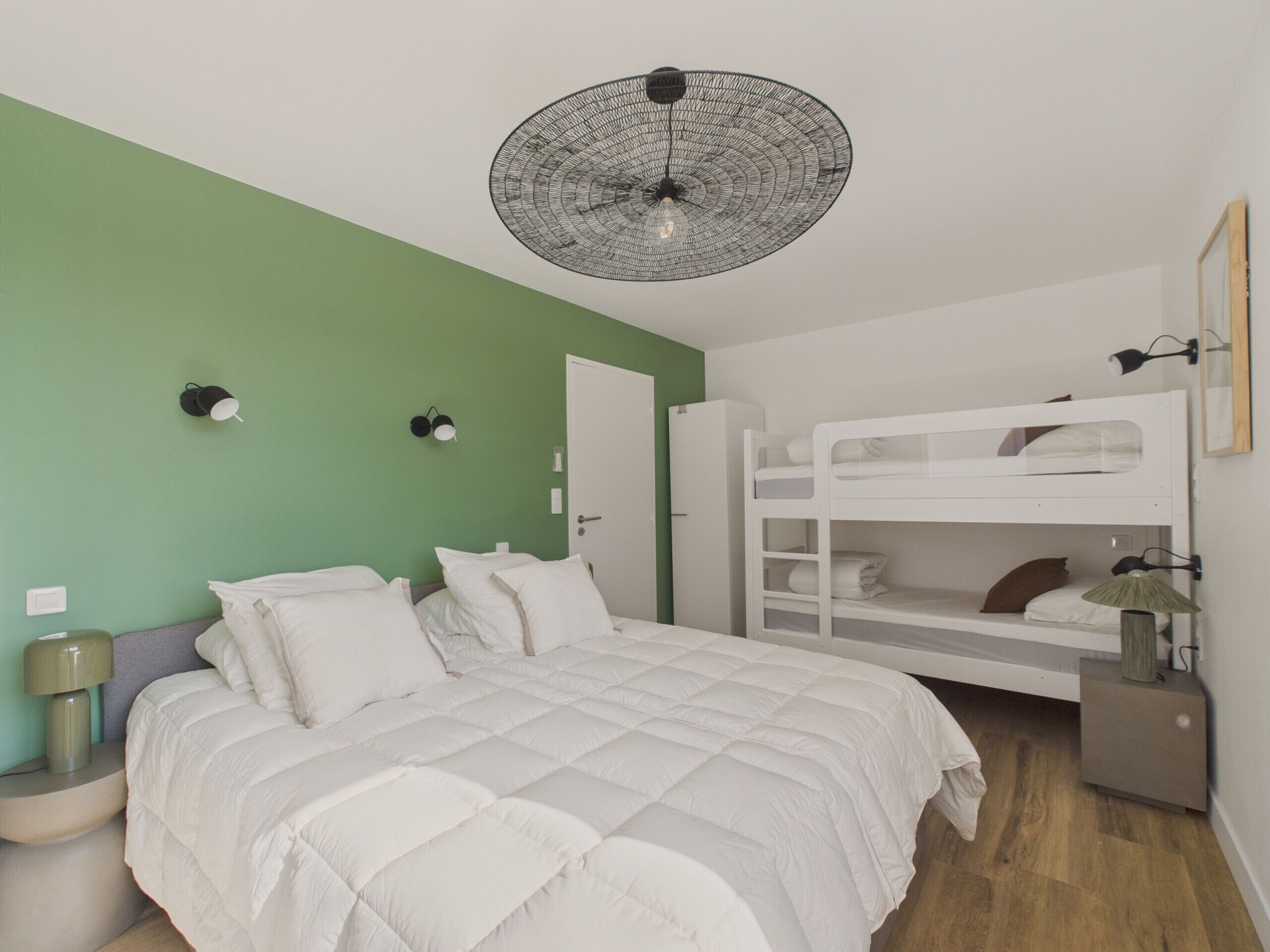 A bedroom with a bunk bed, a white bed with a quilt, a green wall, a round ceiling light, and a bedside table with a lamp.