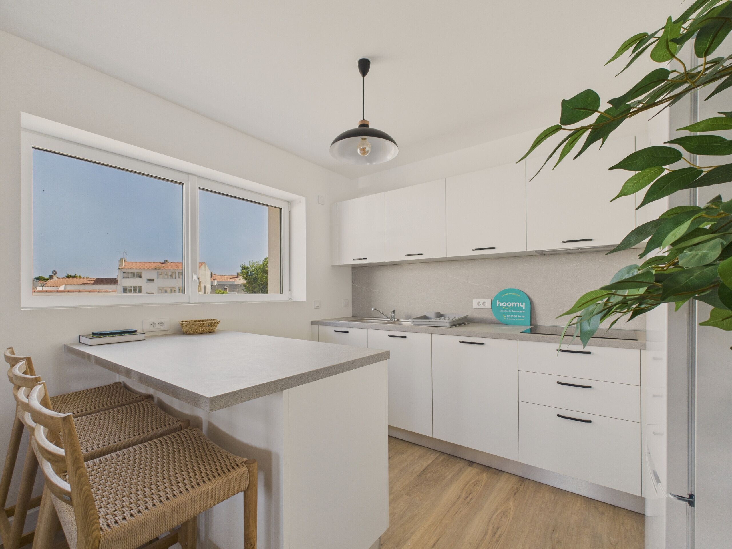 A modern kitchen with white cabinets, a central island, a dining area with wicker chairs, and a view of buildings through a large window.