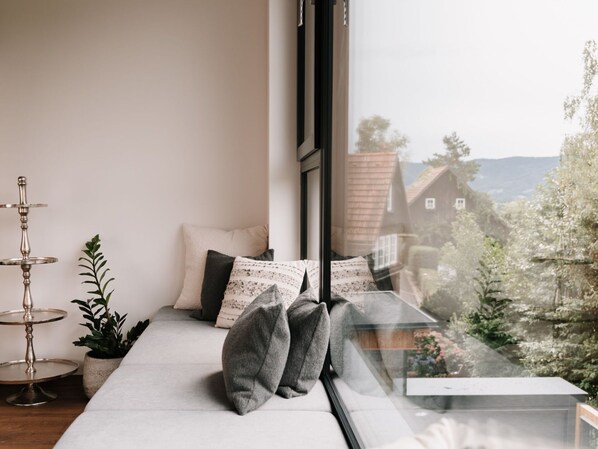 A modern room with a large window, a sofa with patterned cushions, a small plant, and a metal stand.