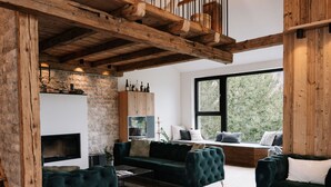 A modern living room with a fireplace, dark green sofas, wooden beams, and a large window offering a view of greenery.