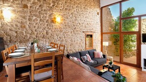 A dining area with a stone wall, wooden table, and chairs. There is a sofa and a window with a view of greenery. - Charming barn in the Matarraña, patio with barbecue and AC - Casa de las Letras (Liedo)