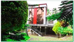 A modern building with a red vertical stripe, surrounded by lush greenery and palm trees. - Green Mount Residency (Devikolam)