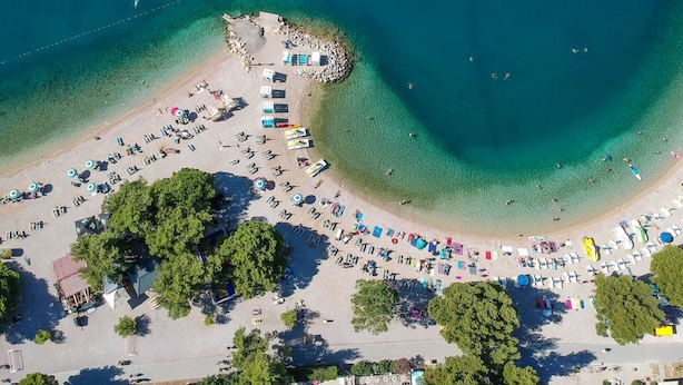 Vue aérienne d’une plage avec des rangées de transats et de parasols, un petit espace rocheux et une eau d’un bleu limpide.