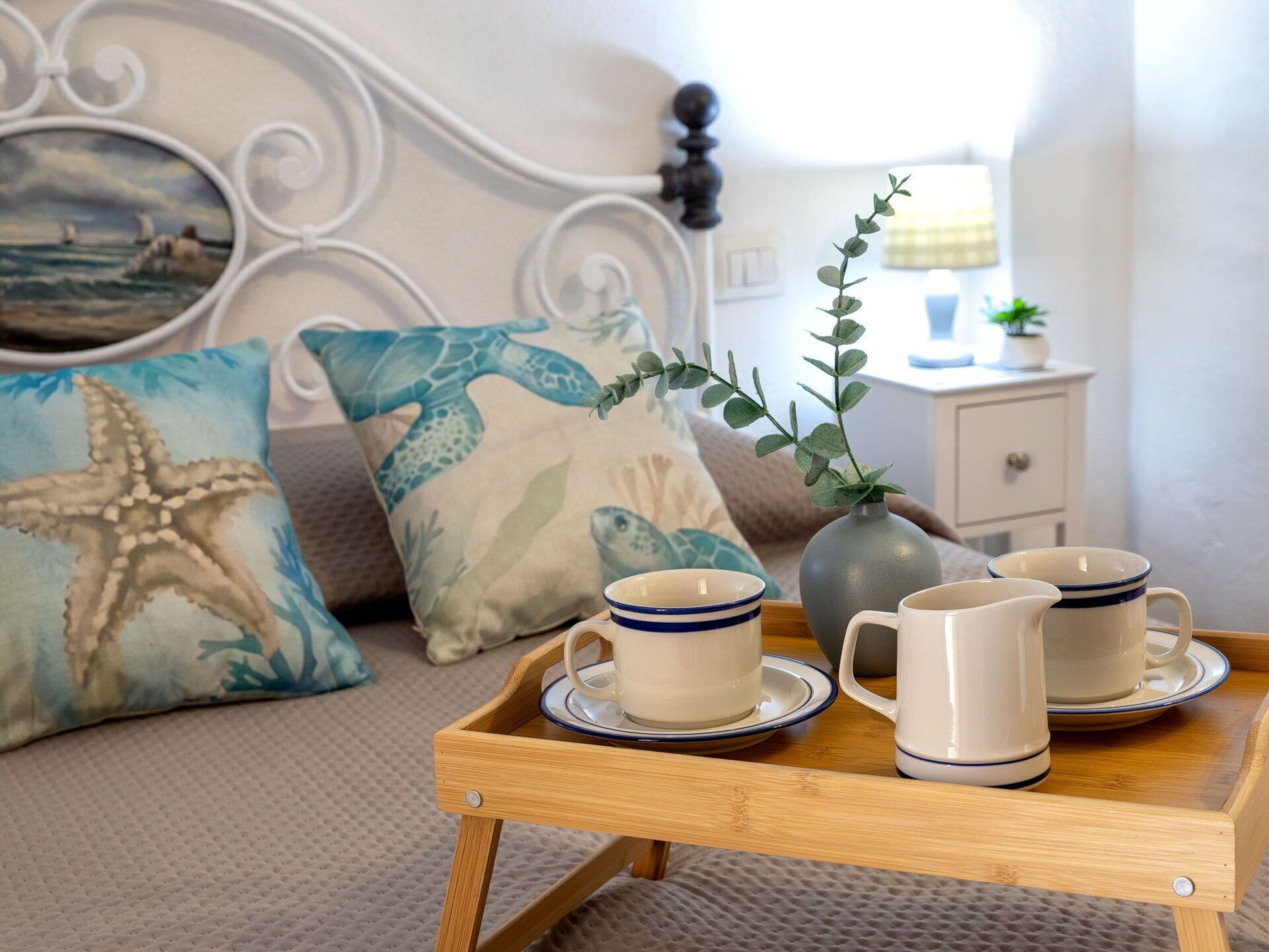 Un lit avec une tête de lit, deux oreillers ornés de motifs représentant des créatures marines, une table de chevet avec un vase et une lampe, et un plateau avec des tasses et une carafe.
