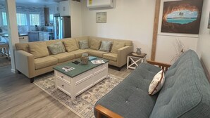 Een woonkamer met een hoekbank, een salontafel en een houten fauteuil. Achtergrond is een keuken met een koelkast en een eethoek.