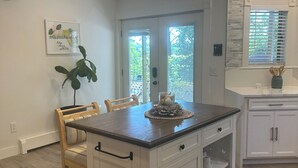 Een keuken met een centraal eiland, witte keukenkastjes en een eethoek met een stoel en tafel.