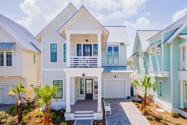A two-story house with a front porch, a garage, and a small garden with palm trees.