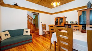 A cozy living room with a green sofa, wooden chairs, a dining table with a white tablecloth, and a staircase leading to another level.
