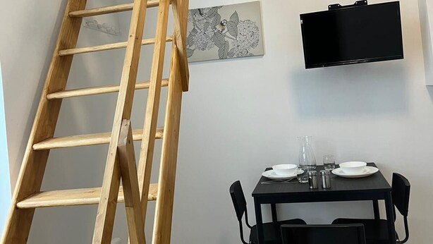 Une cuisine moderne avec une échelle en bois menant à un niveau supérieur, une table à manger noire avec des chaises et un tél éviseur à écran plat fixé au mur.