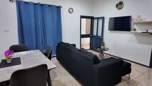 A room with a black sofa, a dining table with chairs, a small round table with a potted plant, a TV mounted on the wall, and a blue door with a mesh screen.