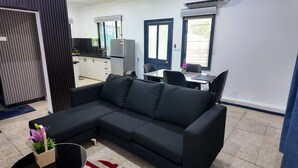A modern living room with a black sectional sofa, a dining table with chairs, a kitchen area, and a small office space.