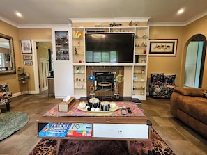 Una sala de estar con un televisor de pantalla plana grande, chimenea, una mesa de centro con juegos de mesa y un sofá.