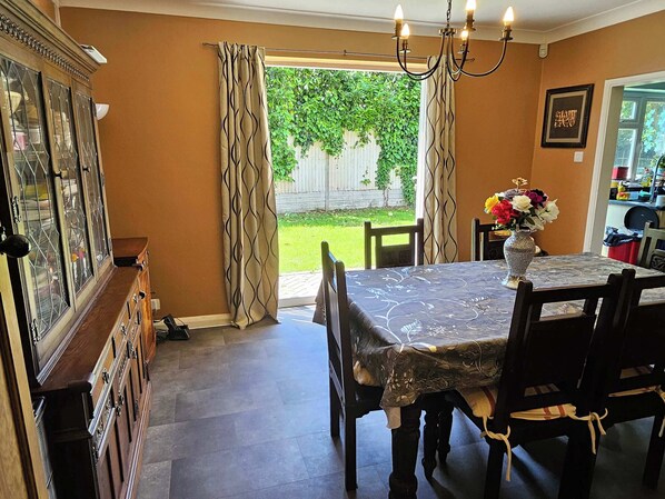 Ein Essbereich mit Tisch, Stühlen, einer Chandelüster, einem Schrank und Blick auf einen Garten durch offene Vorhänge.