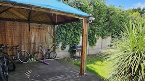 A covered wooden deck with bicycles and a scooter, a barbecue grill, and lush greenery.