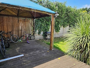 Una terraza cubierta de madera con bicicletas, un scooter, una parrilla para barbacoa y abundante vegetación.