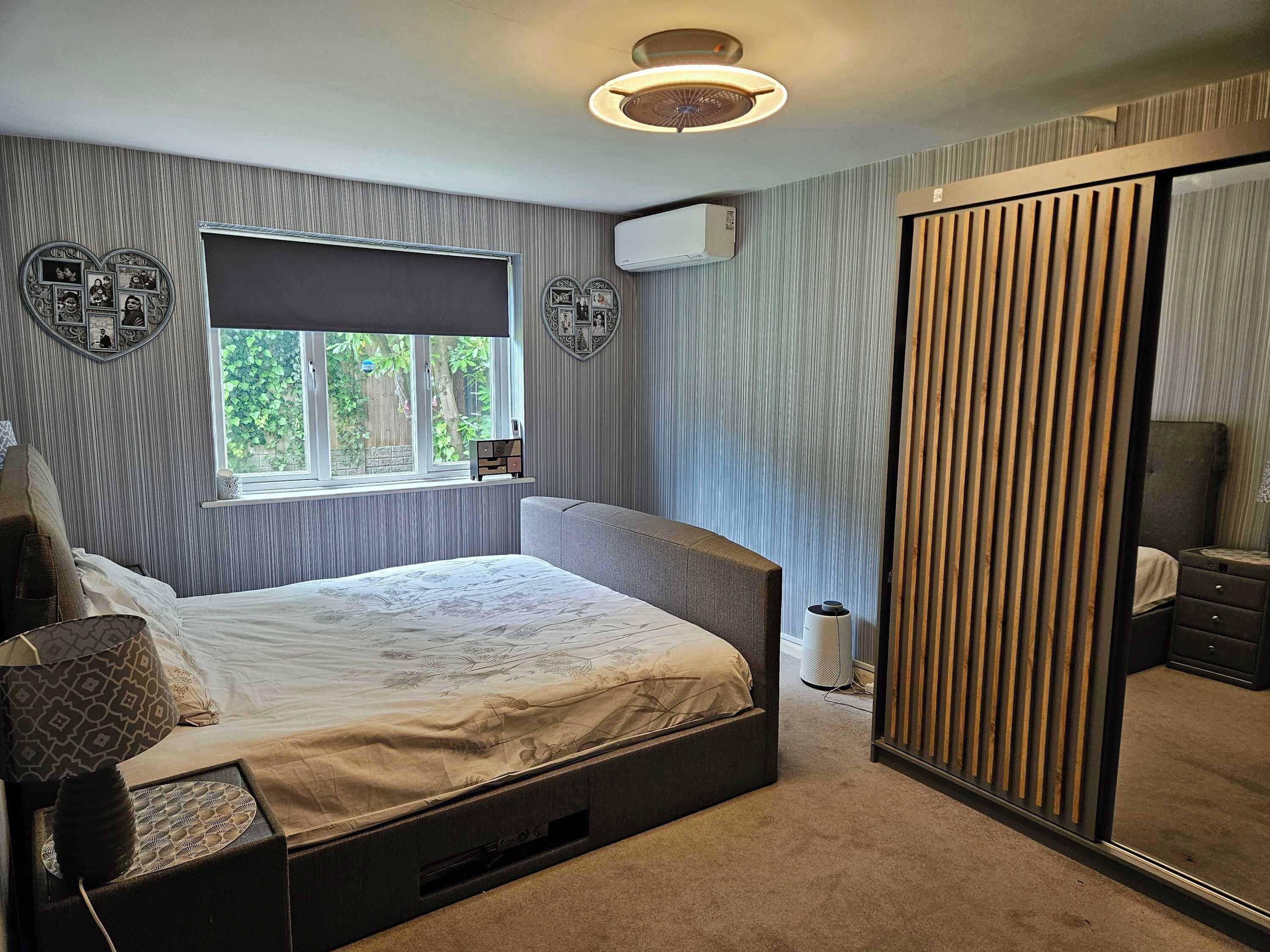 A bedroom with a bed, a window, an air conditioner, a mirror, and a bedside table.