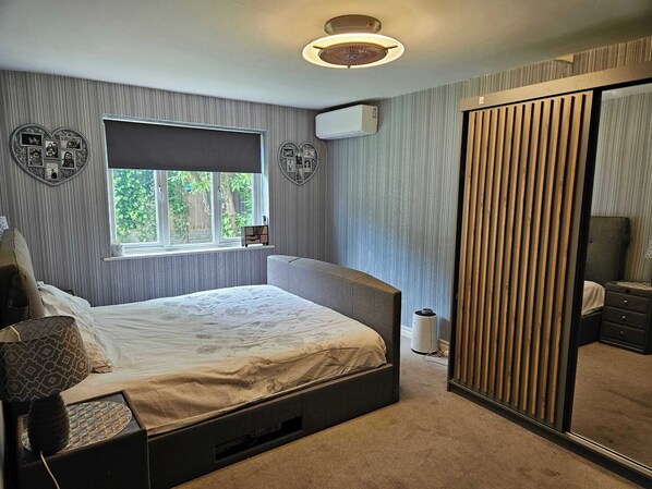 A bedroom with a bed, a window, an air conditioner, a mirror, and a bedside table.