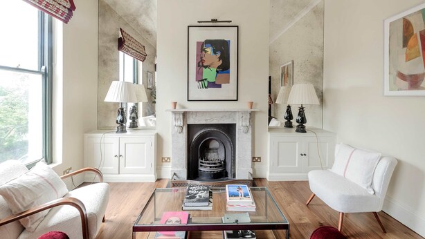 Una sala de estar con chimenea, muebles blancos y una mesa de centro de vidrio con libros.