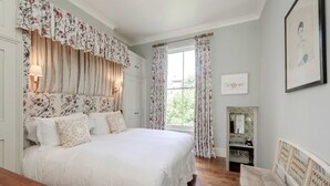 Un dormitorio con una cama grande, cortinas florales, una mesita de noche de madera, un espejo y un cuadro en la pared.