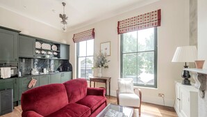 Una sala de estar con un sofá rojo, una mesa de centro de vidrio y un sillón blanco. Hay una zona de cocina con armarios verdes y una encimera de mármol. La habitación tiene ventanas grandes con cortinas rojas y una chimenea con repisa blanca.