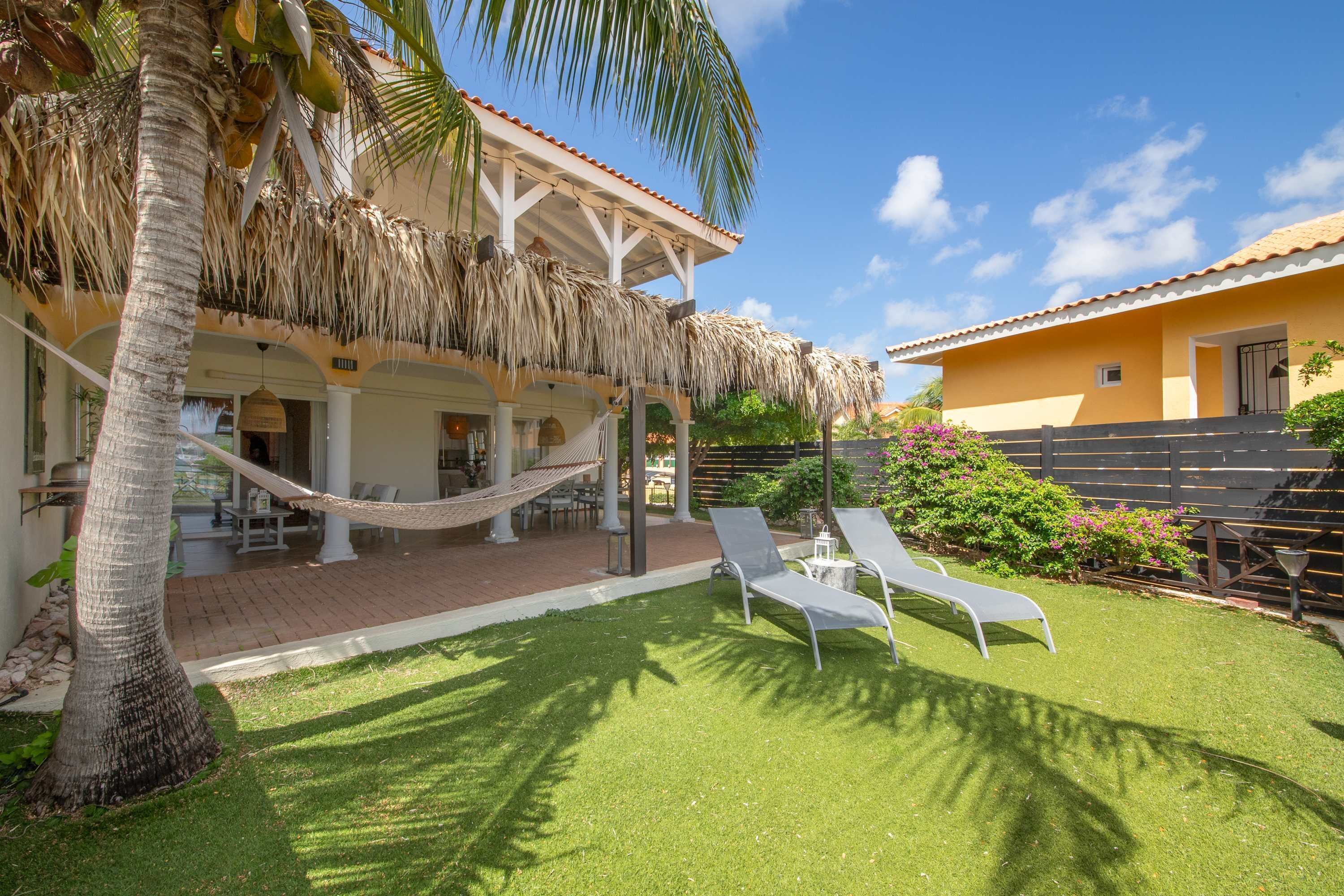 A house with a thatched roof patio, a hammock, and a lawn with lounge chairs.