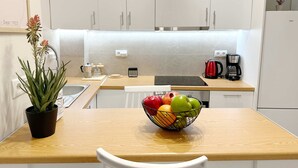 Une cuisine moderne avec des meubles blancs, un plan de travail en bois, un évier, une bouilloire, une machine à café et un panier de fruits.