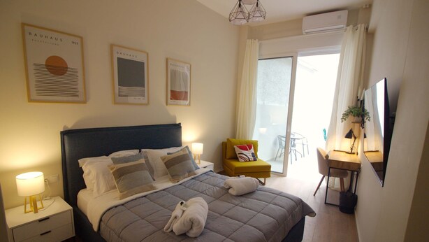 A neatly made bed with pillows, a bedside table with a lamp, and framed artwork on the wall. - Secret Garden by Acropolis - Athens Central Gem (Athens)