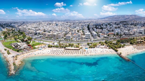 Vue aérienne d’une ville côtière avec une plage, des bâtiments et une chaîne de montagnes en arrière-plan.
