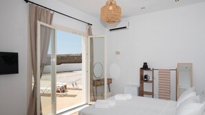 A hotel room with a bed, a desk, a chair, a mirror, a hanging light fixture, and a view of the beach through the door. - Adyton Lux Suite Mykonos Private Pool Experience (Mykonos)