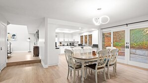 Una cocina moderna con armarios blancos, un comedor con mesa y sillas de madera, y un amplio ventanal corredizo que da a un balcón con vistas a la vegetación.