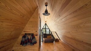 Interior de una cabaña de madera con una cama en el altillo, una silla y una lámpara colgante.